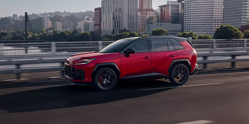 Red Toyota RAV4 driving on a bridge with a city skyline in the background.
