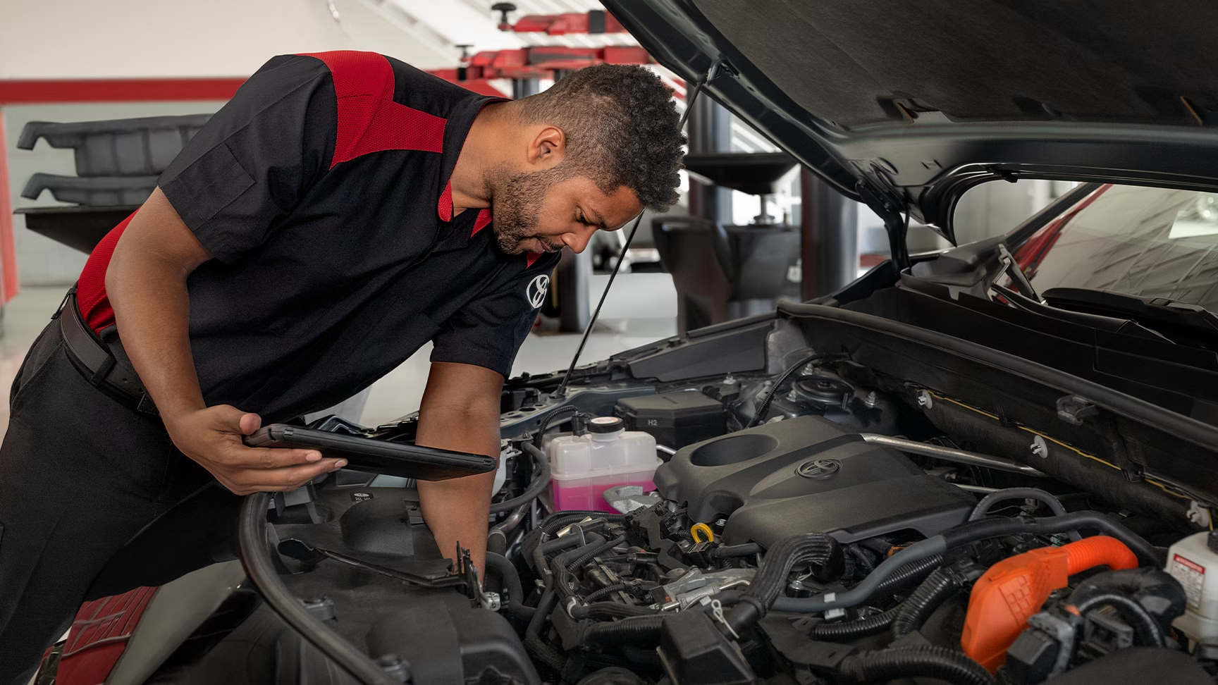 A Toyota mechanic inspecting a car engine bay while holding a digital tablet.